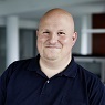 Smiling man with a black polo shirt and a bald head.