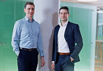 Two men dressed in shirt and suit in front of gray pillar with green background.