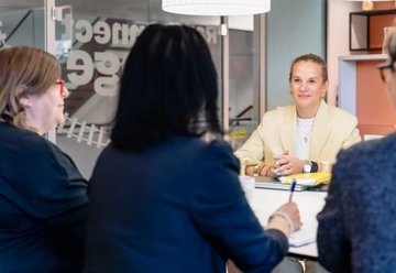 Four women in a meeting room; one woman in a light blazer sits opposite the others, appearing attentive and engaged.