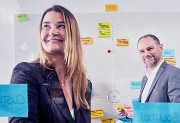 A smiling woman as well as a smiling man in a suit in front of a transparent wall with notes and post-its.