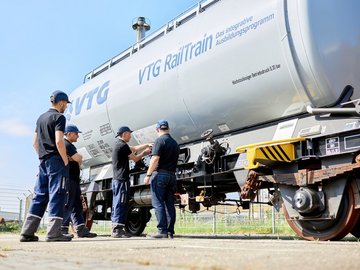 Many employees in protective clothing in front of a gray VTG tank car.