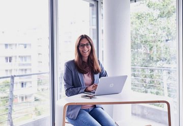 Smiling woman in front of her laptop with window panes in background.