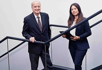 An older smiling man in a suit and a smiling younger woman standing on a staircase.