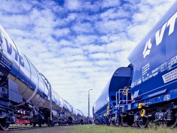 Silvery VTG chemical tank car next to blue VTG standard freight car under blue sky